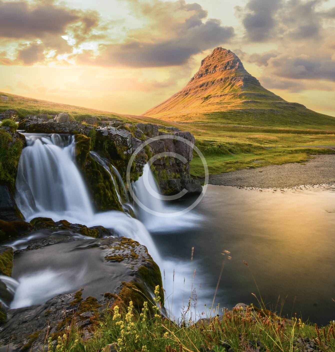 A waterfall in the middle of a field with mountains behind it.